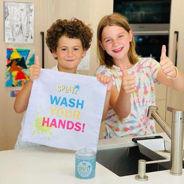 One child holding a splatz "wash your hands" hand towel, with another child giving a thumbs up