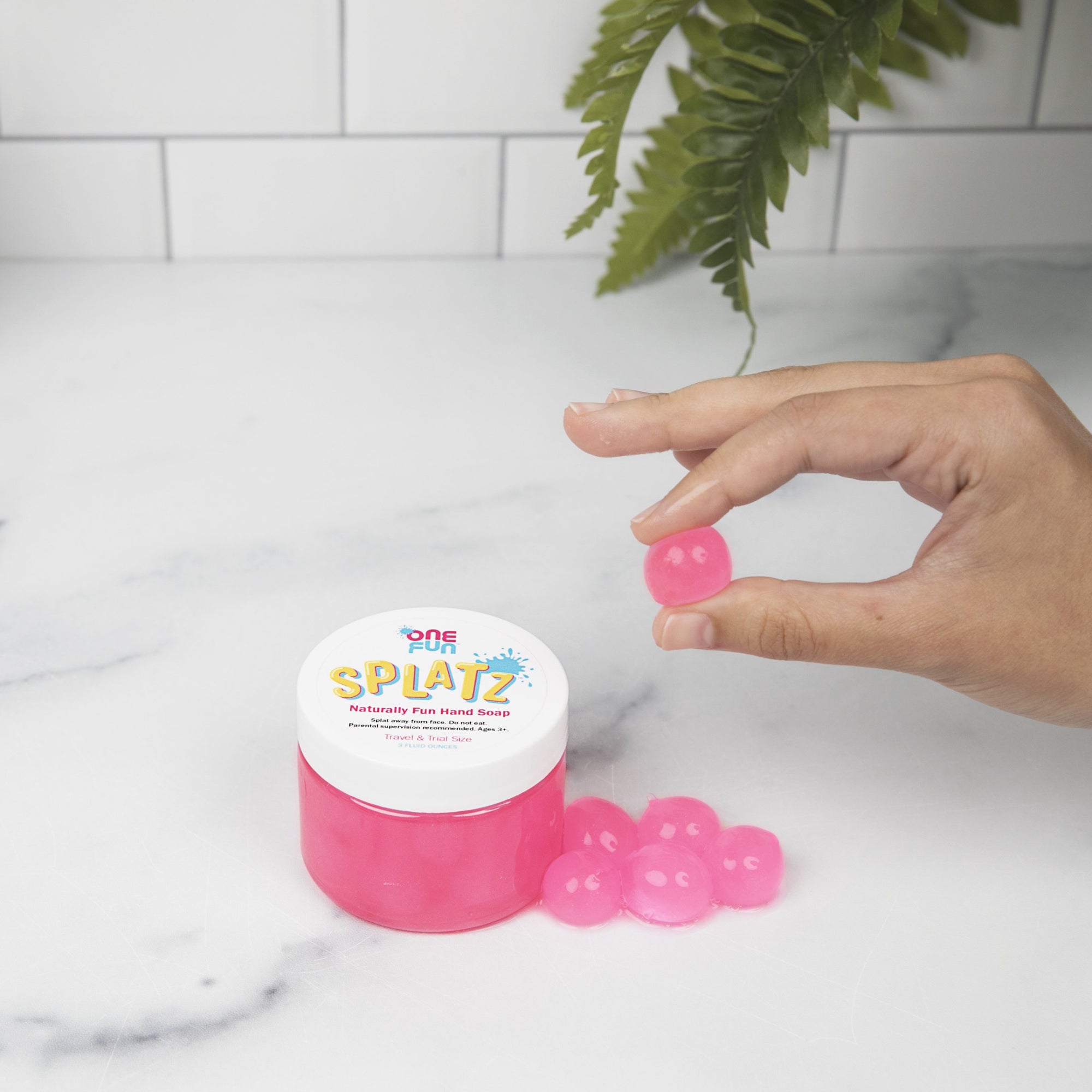On a bathroom counter. One 3oz jar of pink bursting soap bubbles. A hand holding one pink bursting soap bubble over the jar